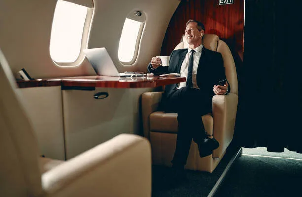 A man sits in a leather chair inside a private jet, working on a laptop.