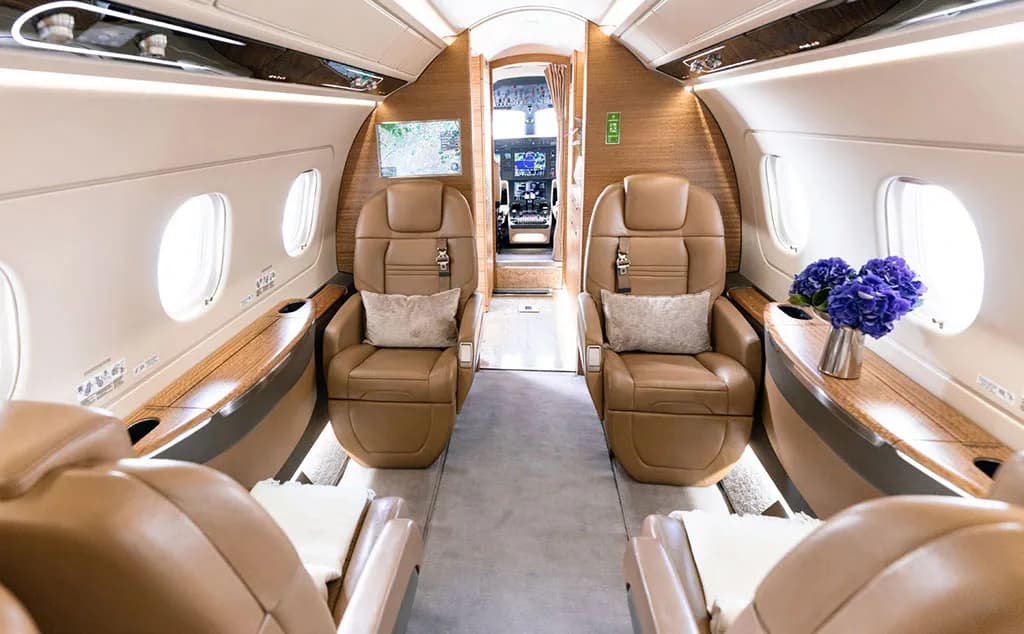 A view down the aisle of a business jet cabin with tan leather seats and a vase of blue flowers.