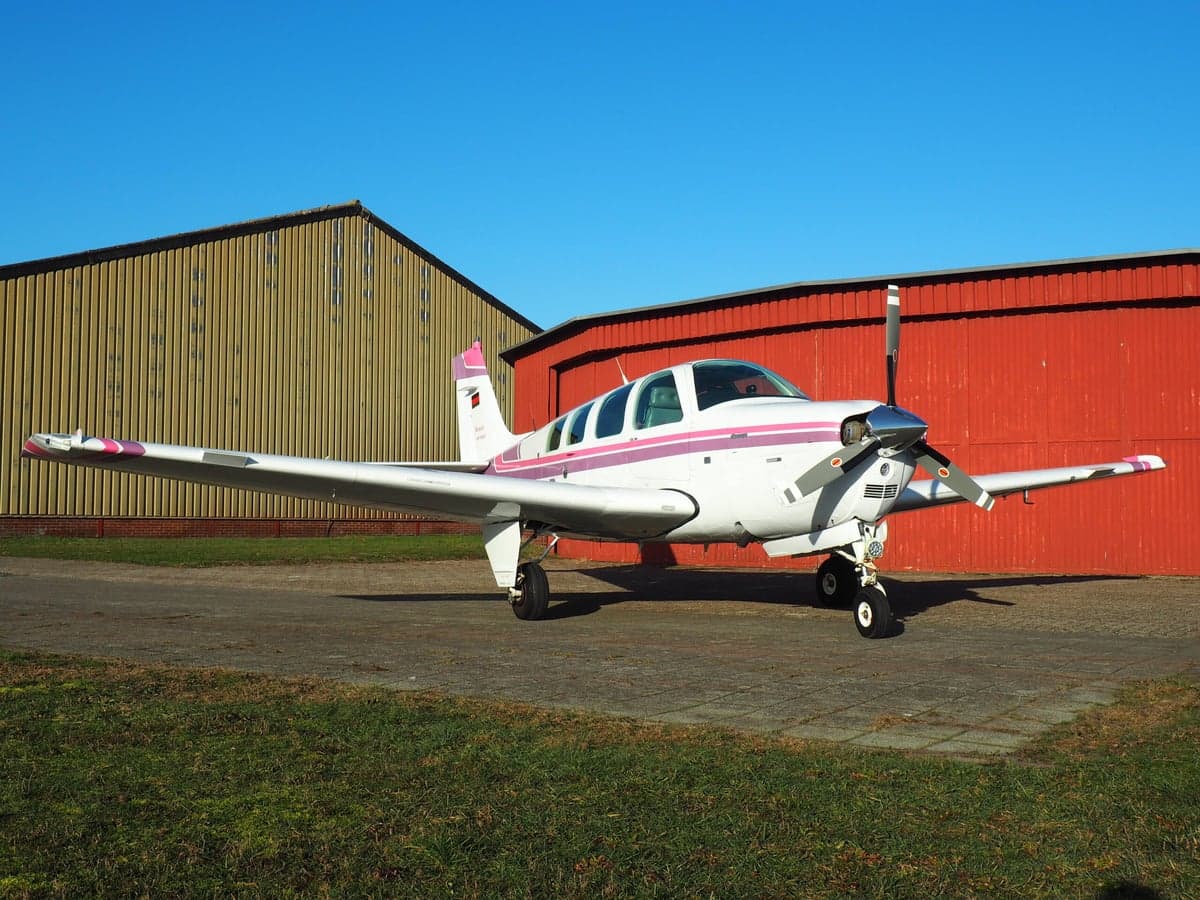 A white and pink Beechcraft Bonanza parked on the tarmac in front of a hangar.