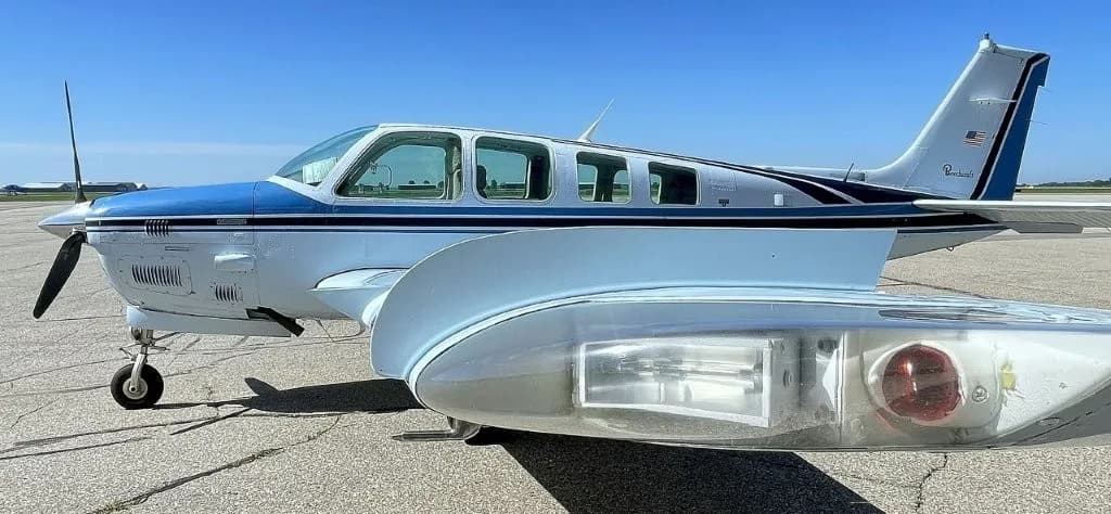 Side profile of a white and blue Beechcraft Bonanza aircraft on the tarmac.