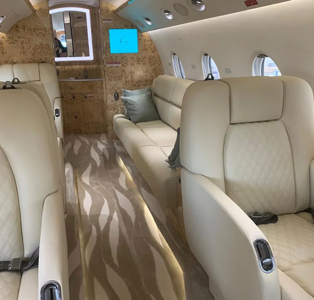 A wide shot of the private jet cabin interior, featuring cream leather seats, a sofa, and a patterned carpet leading to a mirrored wall.