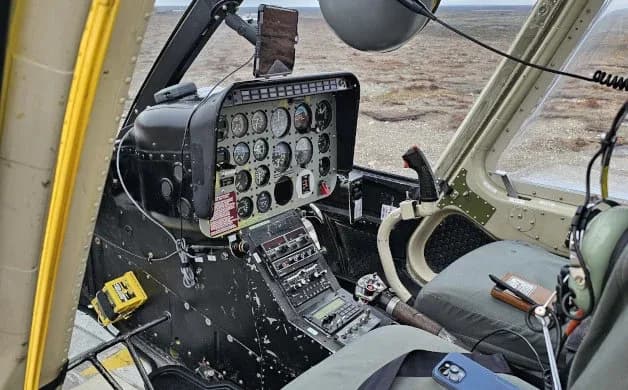 The cockpit of a helicopter features a detailed instrument panel, cyclic, collective, and worn pilot's seat with a headset.