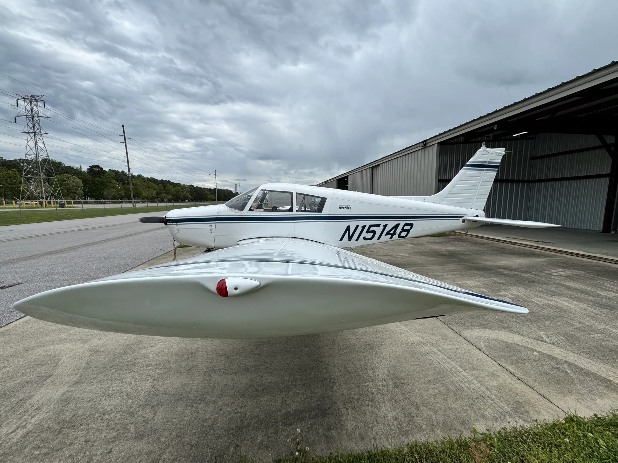 A white single-engine aircraft, N15148, with dark blue stripes, parked on concrete next to a hangar under a cloudy sky.