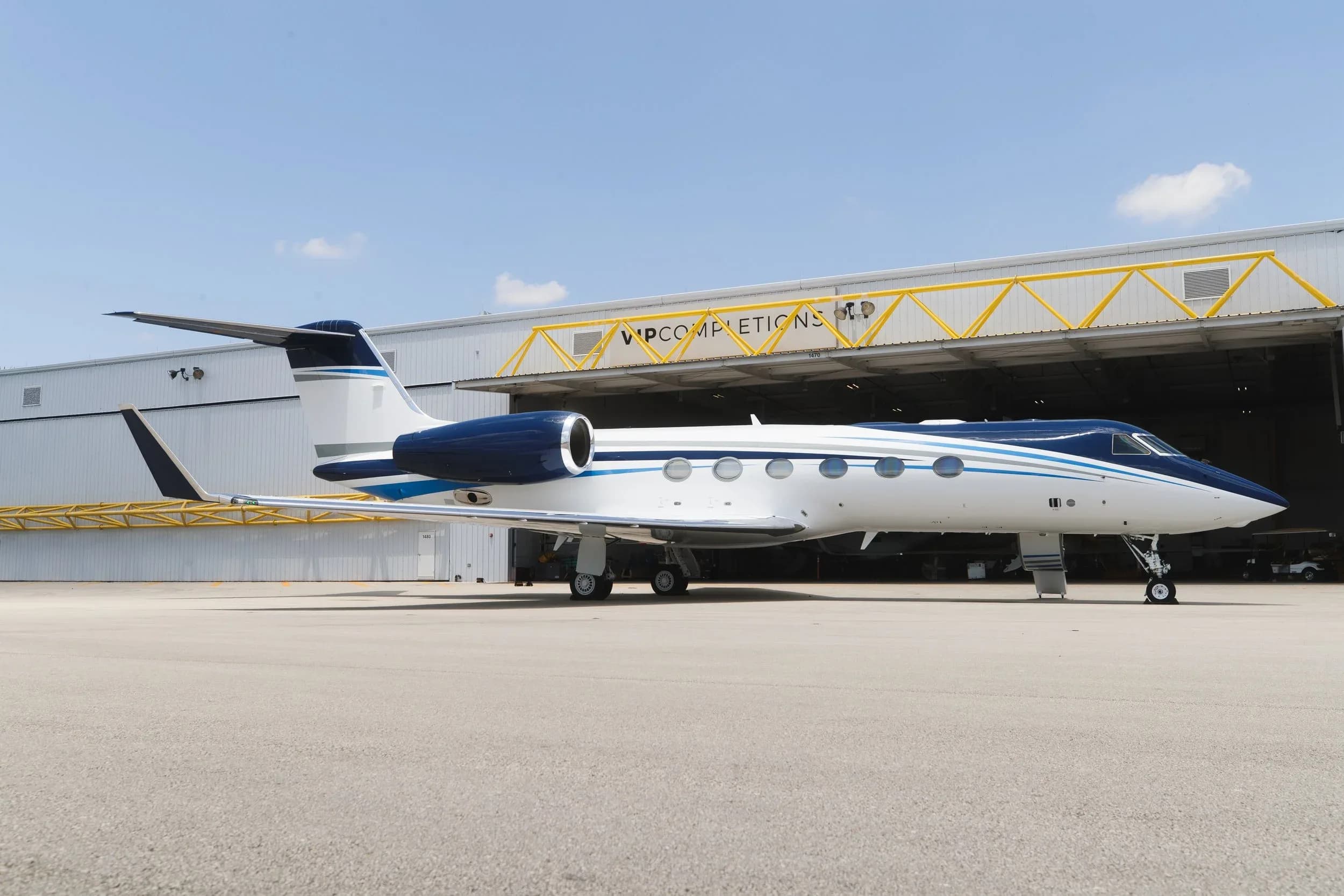 A side view of a blue and white Gulfstream G550 business jet parked on an airfield in front of a hangar.