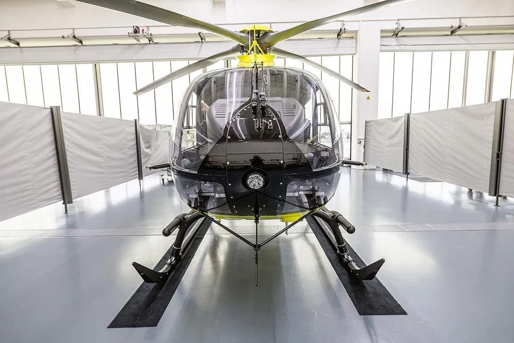A bright yellow Airbus H135 helicopter is viewed head-on in a hangar, showing its large transparent cockpit.