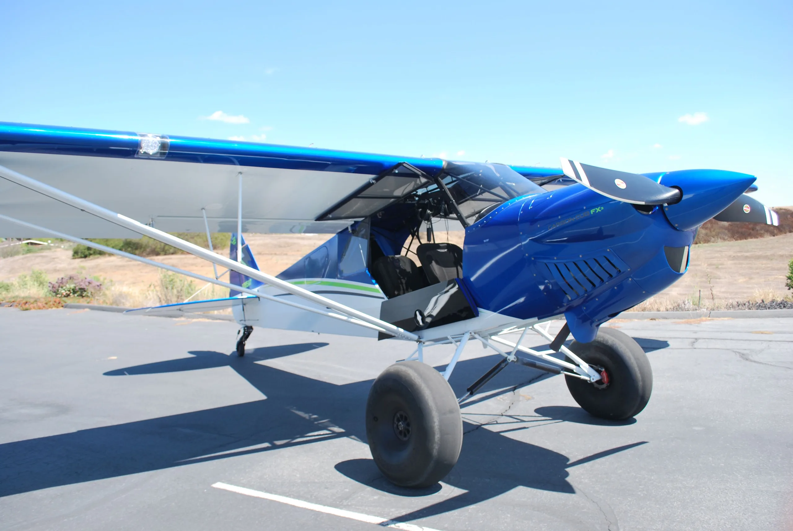 A three-quarter front view of a blue and white Carbon Cub FX3 aircraft with its door open, parked on an asphalt ramp.