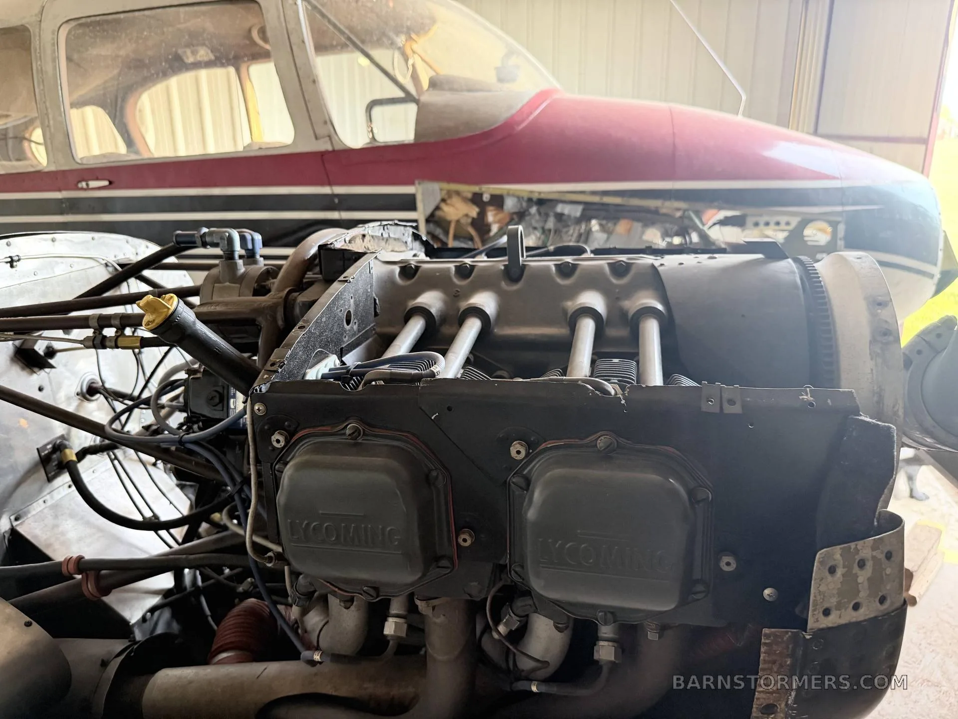 A Lycoming aircraft engine with its cowling open, showing the cylinders and components, with the red and white fuselage in the background.