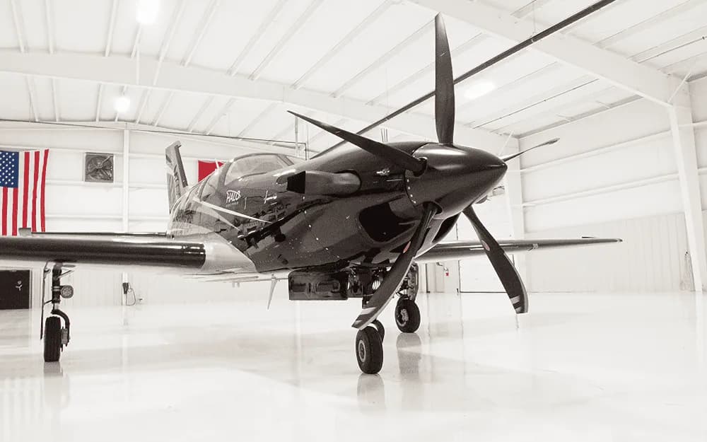 A front view of the black and silver Piper M600/SLS aircraft with its propeller, parked inside a hangar with an American flag in the background.