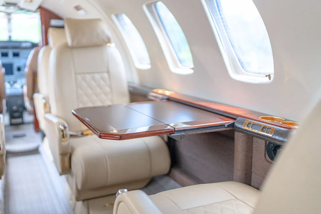 A close-up of a light beige leather cabin seat and a polished wooden fold-out table in a private jet.