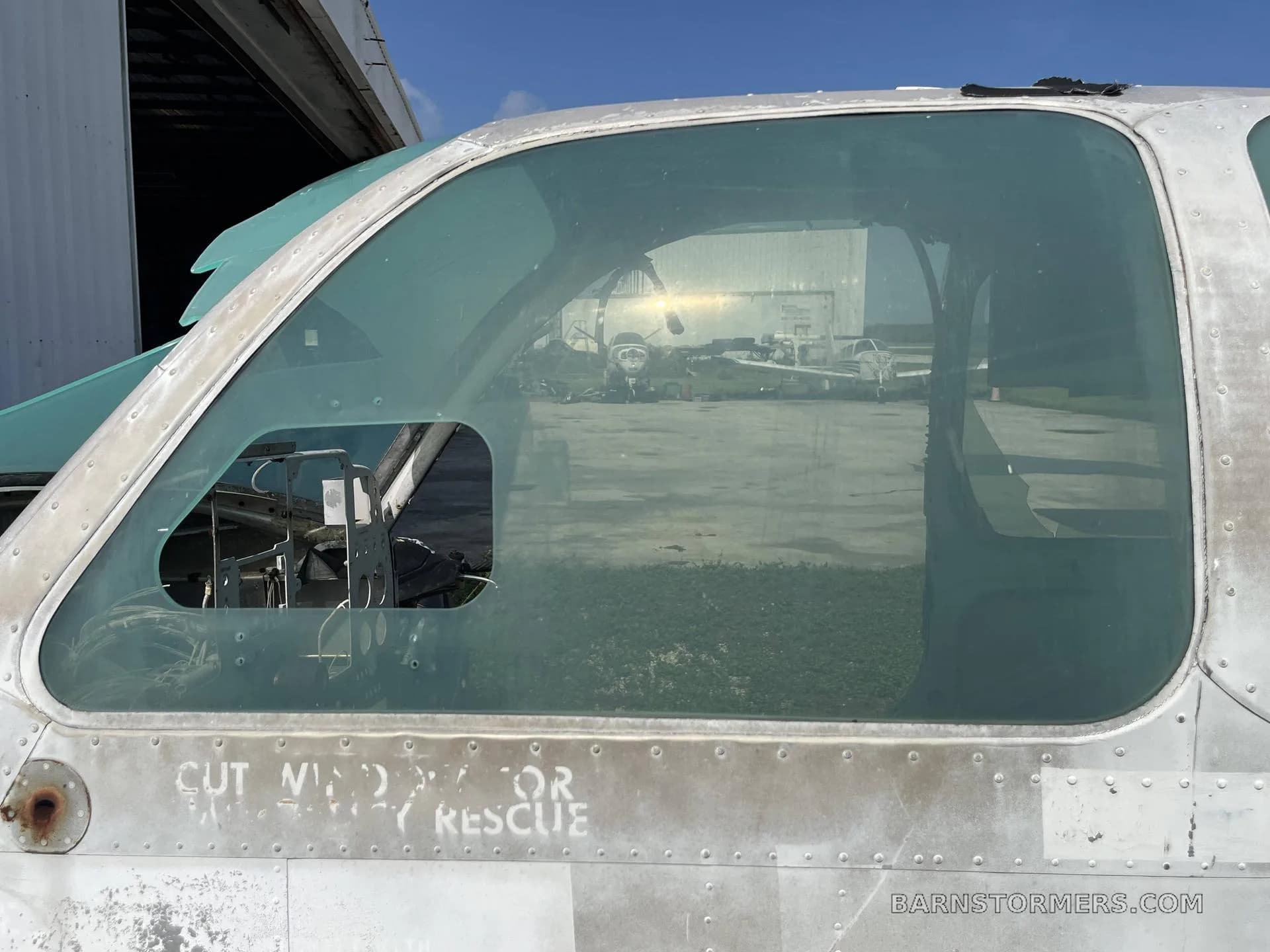A close-up of the weathered cockpit window of an aircraft fuselage, reflecting other aircraft in the distance.