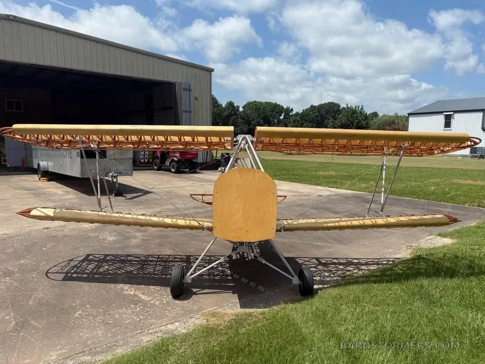 A front view of a partially assembled wooden biplane aircraft frame with landing gear, parked outside a hangar.