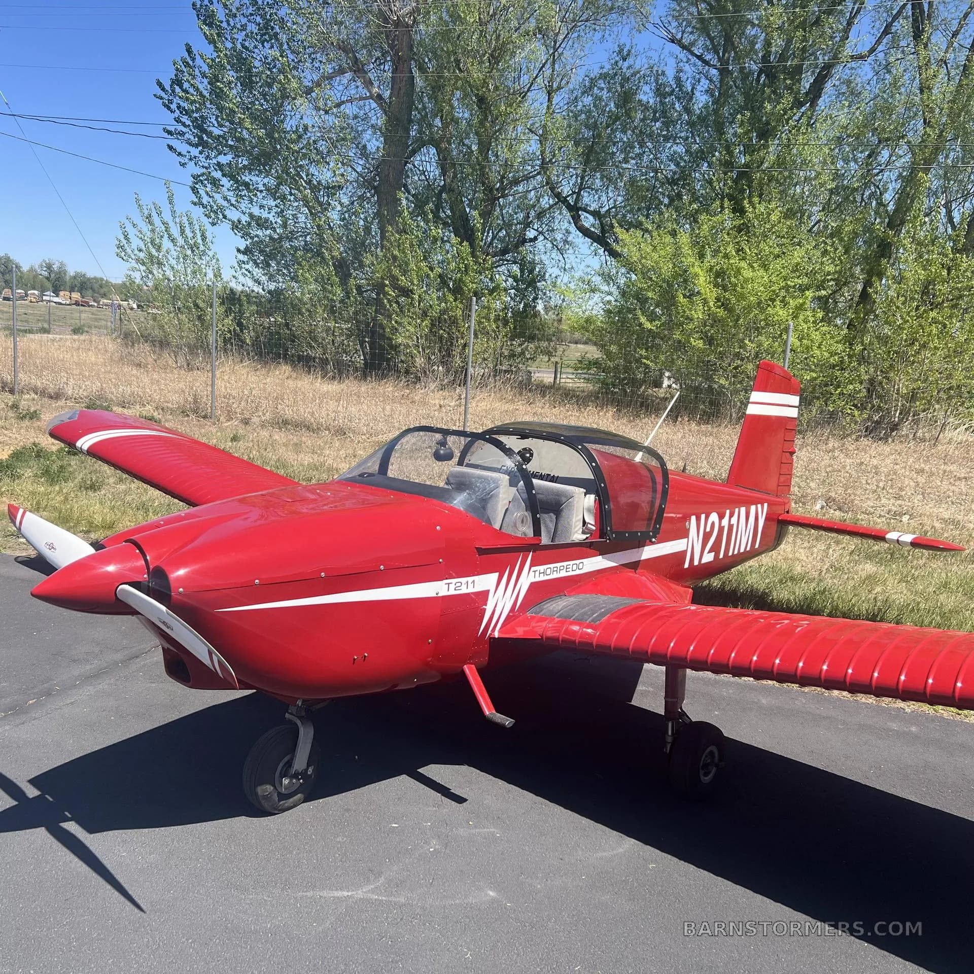 A red Thorp T211 Torpedo aircraft with white stripes and registration N211MY, parked on an asphalt surface with trees in the background.