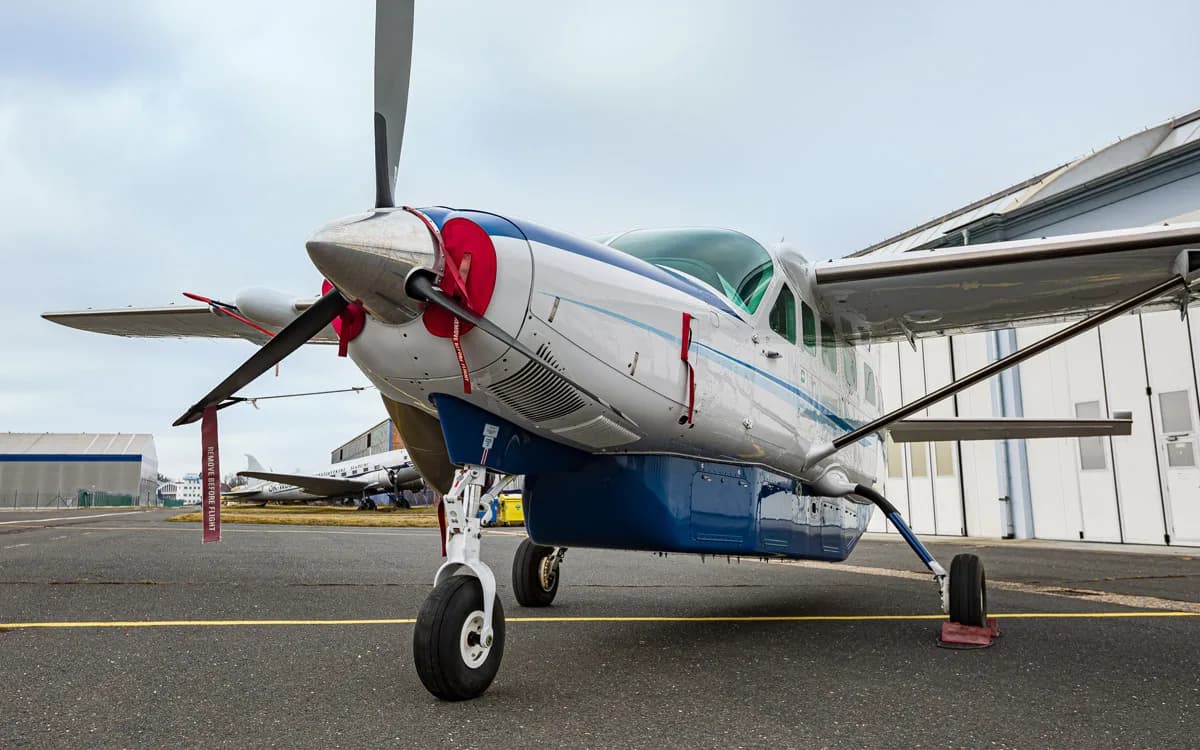 A white and blue Cessna Caravan turboprop aircraft with red 'REMOVE BEFORE FLIGHT' tags, parked on an airport apron.