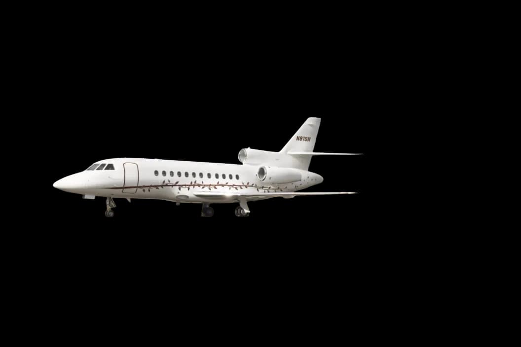 A white Dassault Falcon business jet with a brown vine-like livery detail on the fuselage, seen from the side on the ground.