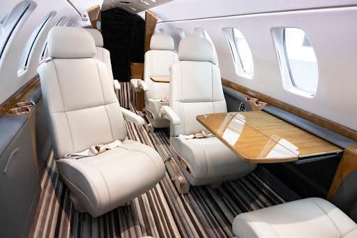 The luxurious cabin interior of the Cessna Citation CJ3+ with light cream leather seats, a striped floor, and a wooden fold-out table.