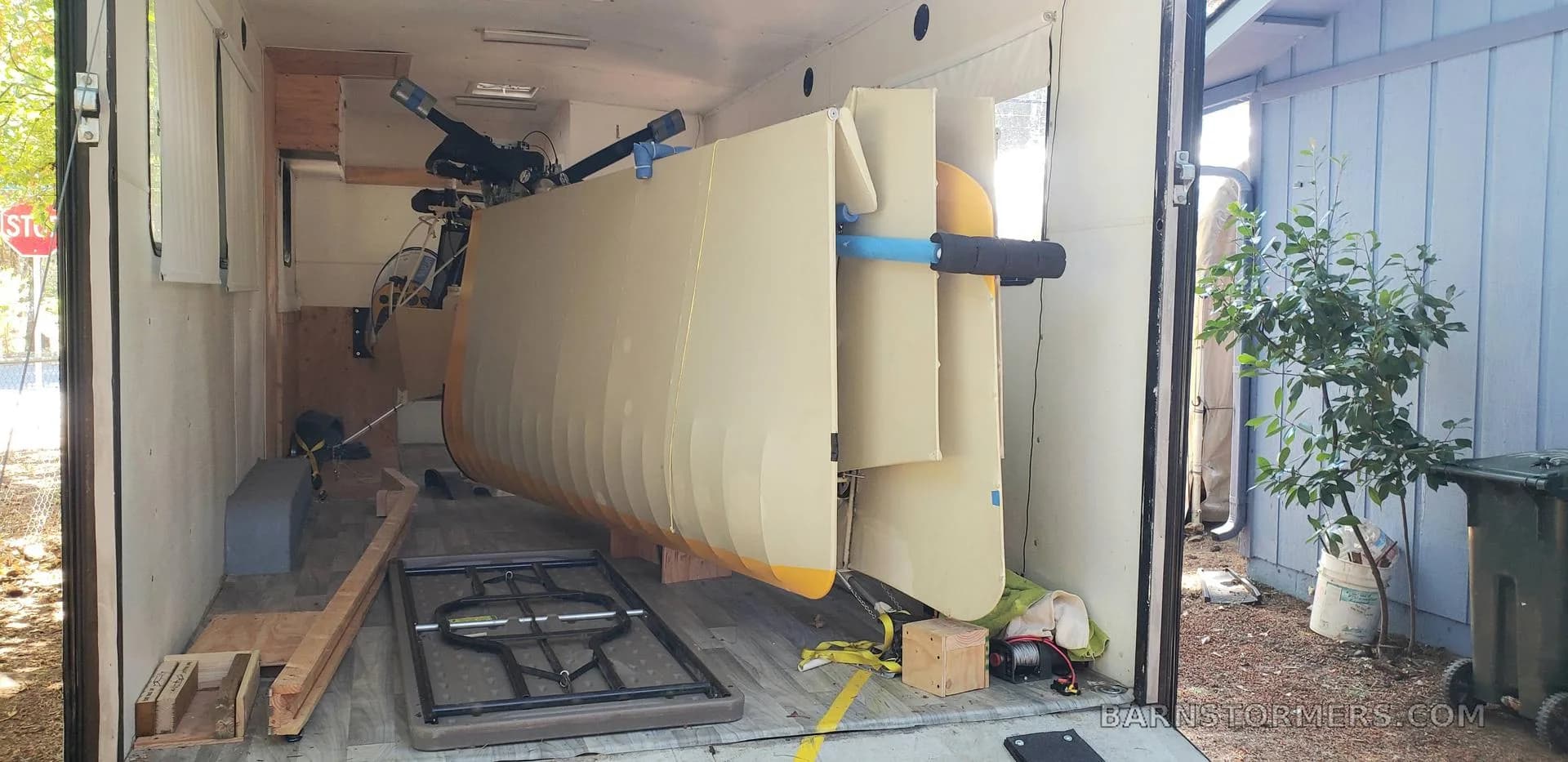 The yellow and white fuselage of an ultralight aircraft is partially loaded into a white enclosed trailer.
