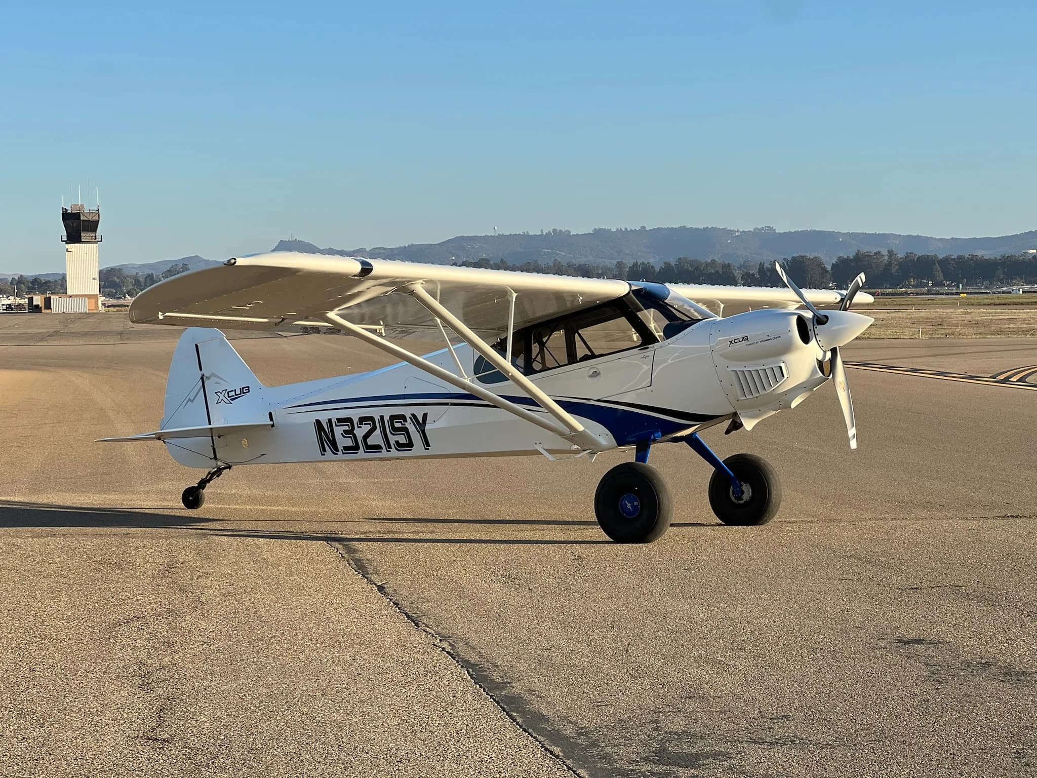 A white X-Cub aircraft with blue stripes and registration N321SY parked on an airfield with a control tower and mountains in the distance.
