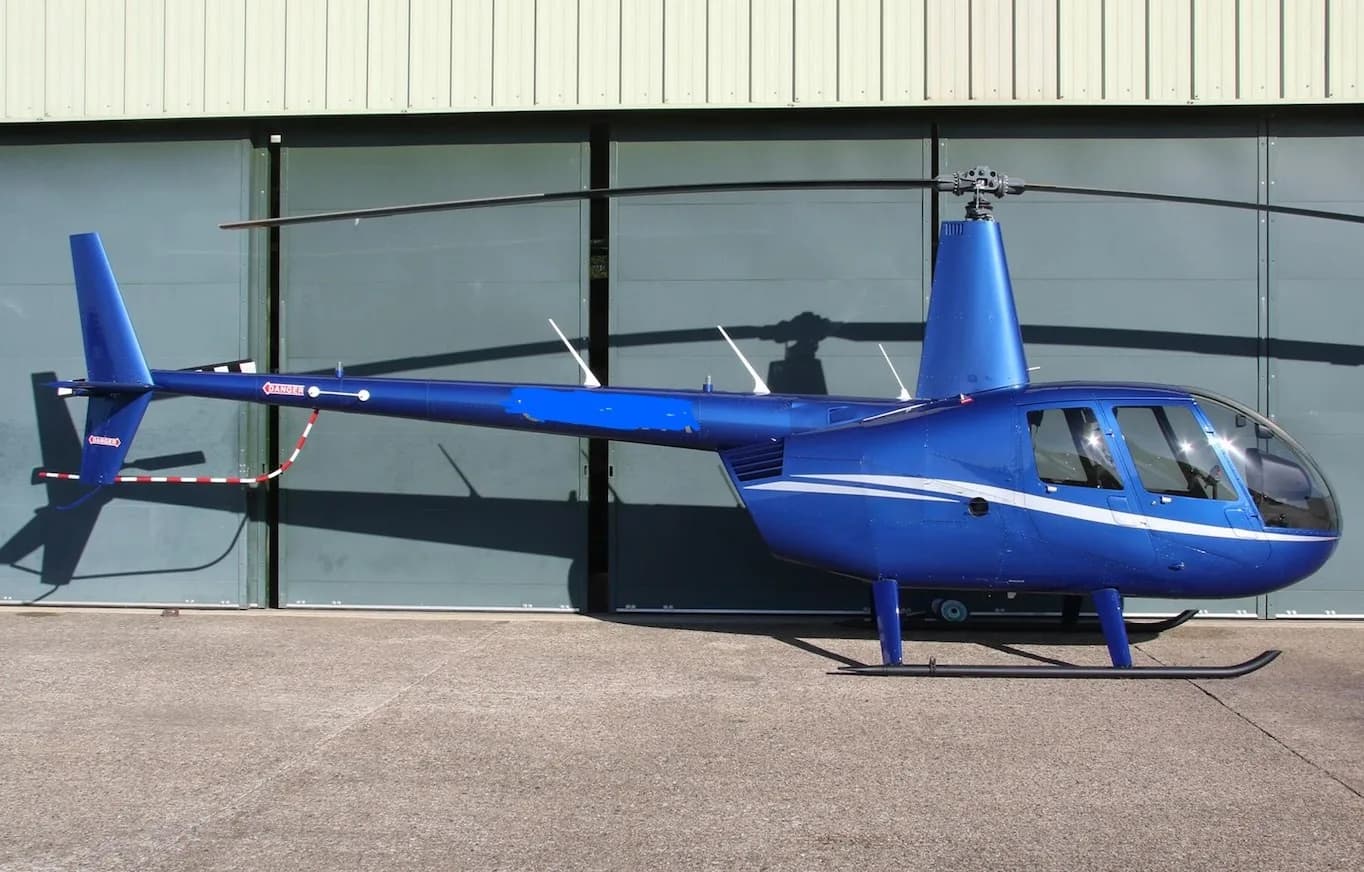 A blue Robinson R44 helicopter with white stripes parked on a tarmac in front of a hangar.