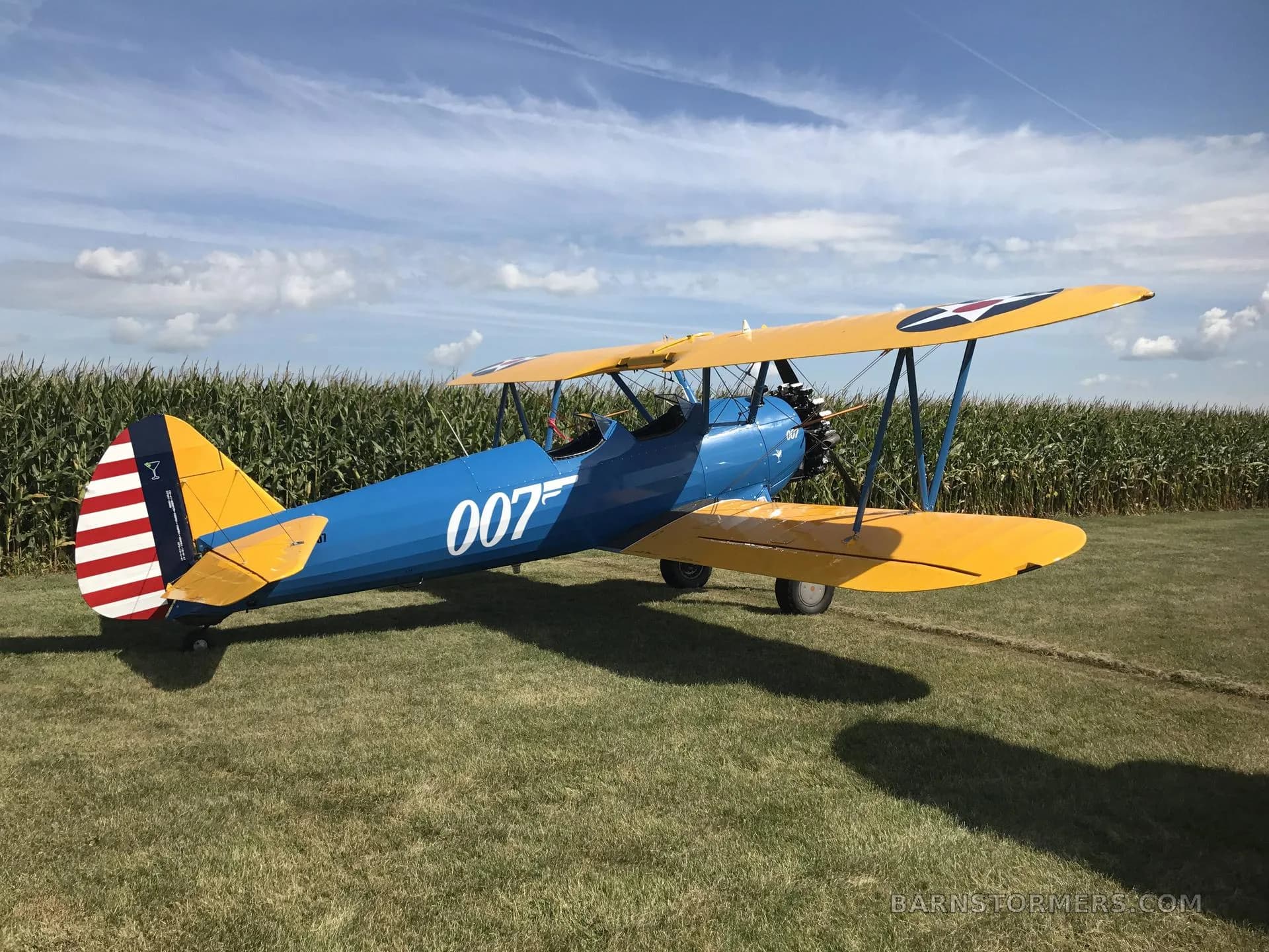 A blue and yellow biplane with "007" on its side is parked on a grassy field next to a cornfield under a partly cloudy sky.
