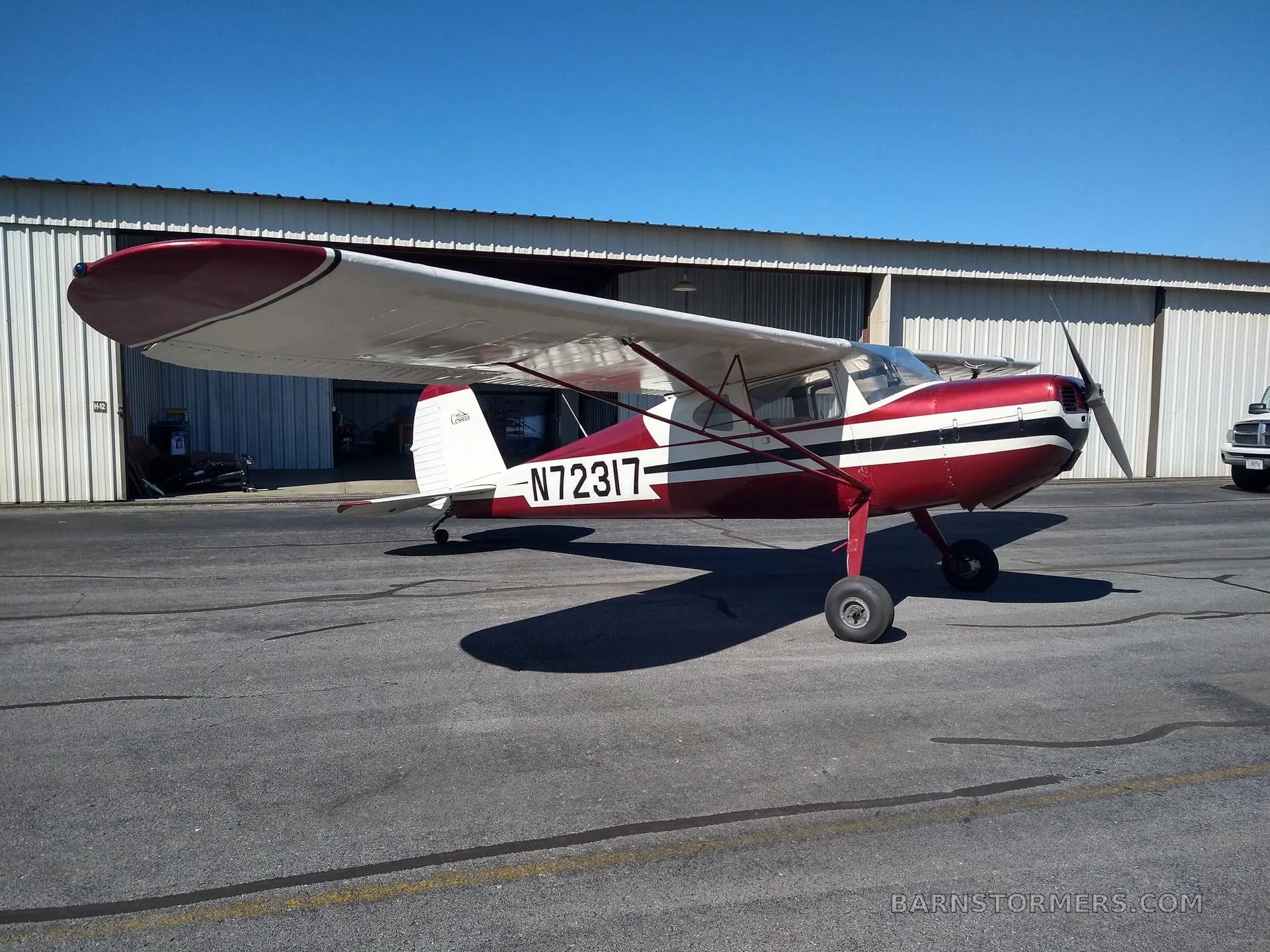 A red and white Cessna 140 aircraft (N72317) is parked on an asphalt tarmac in front of a hangar on a sunny day.