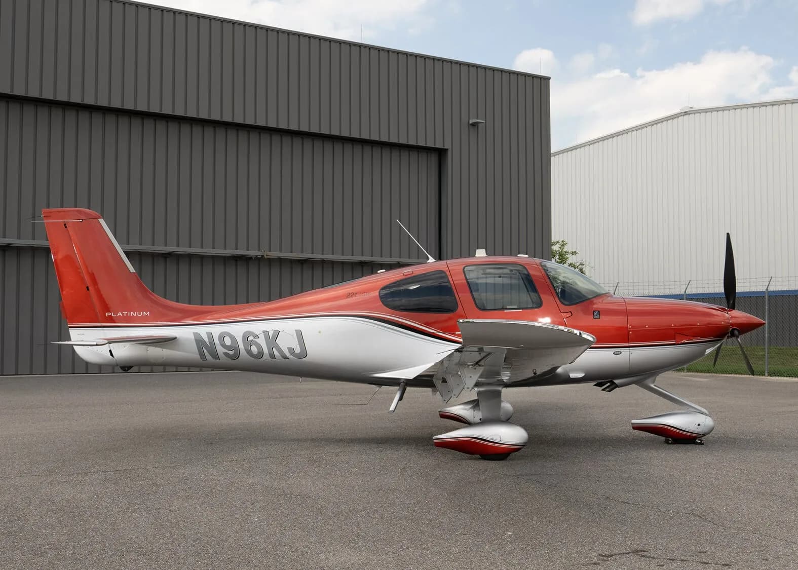 A red and silver Cirrus SR22T aircraft, registration N96KJ, parked on an asphalt apron in front of a hangar under a blue sky.
