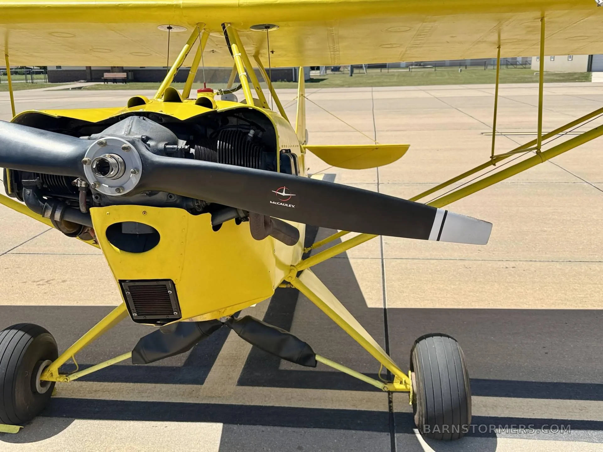 A close-up front view of a yellow biplane's engine, propeller, and landing gear on an airfield.