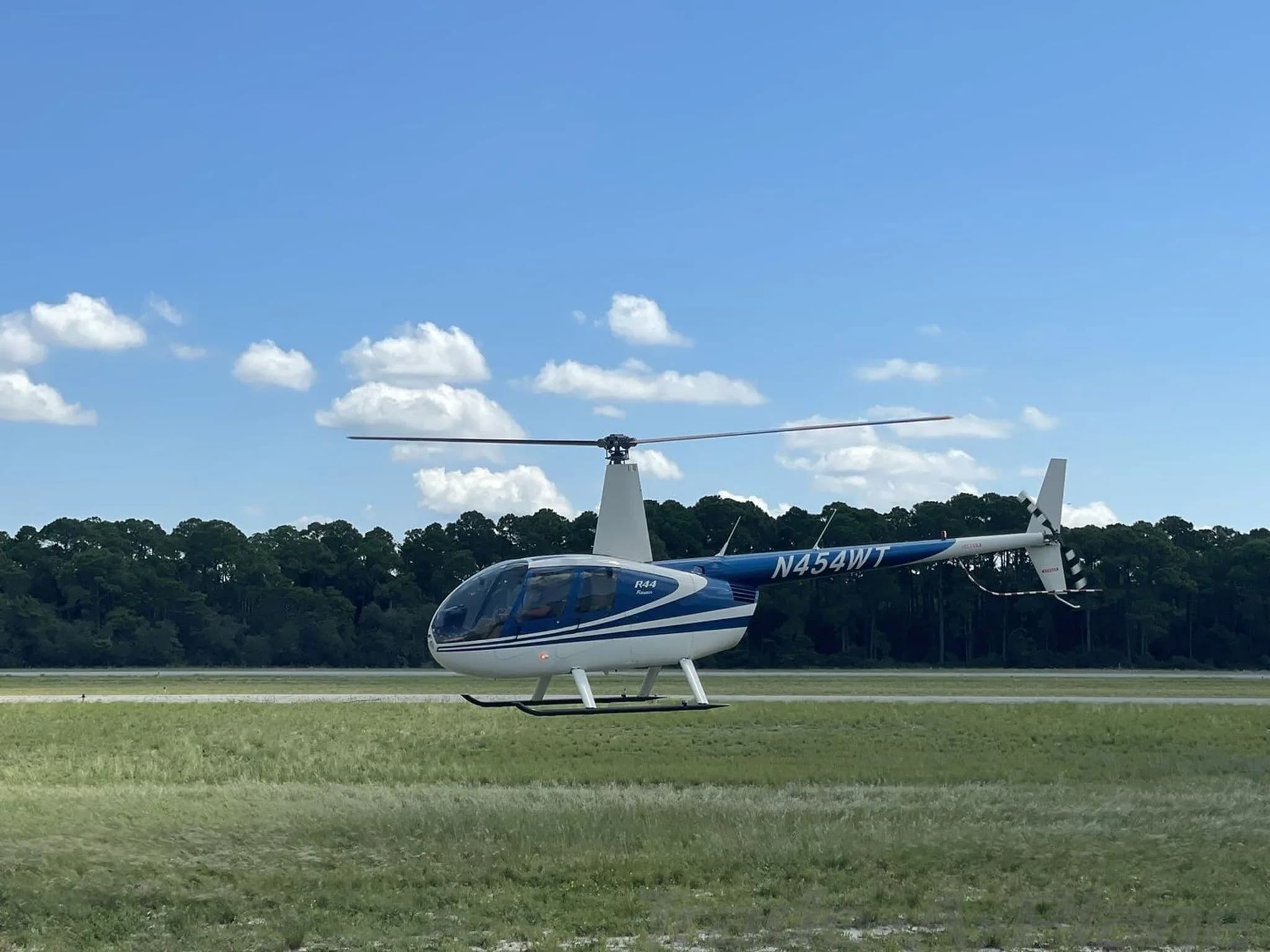 A blue and white Robinson R44 Raven helicopter with registration N454WT hovers over a grassy field under a blue sky.