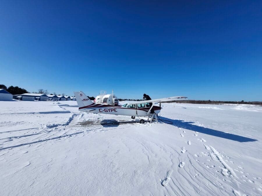 A white and red Cessna 172 (C-GYPE) is parked in a snowy field with a person near its wing.