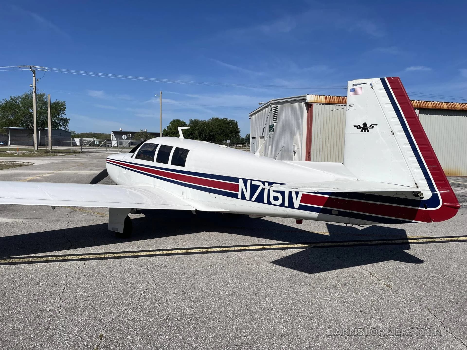 Rear-side view of a white single-engine propeller aircraft with red and blue stripes on the tail and fuselage, parked on an asphalt ramp.