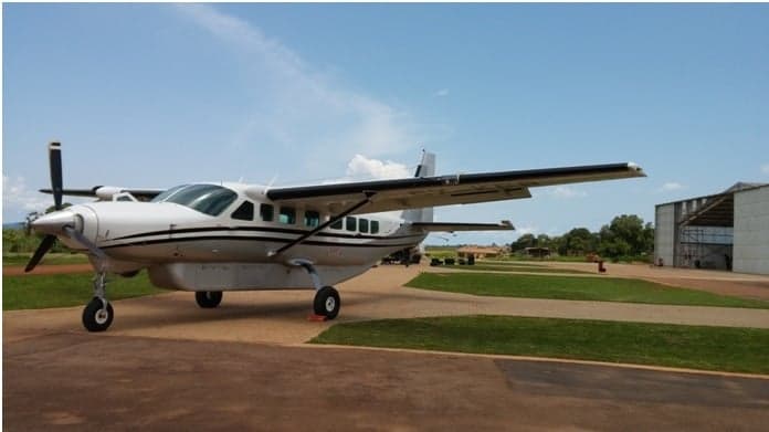 A white twin-engine propeller aircraft with black stripes is parked on an asphalt apron next to a hangar under a blue sky.