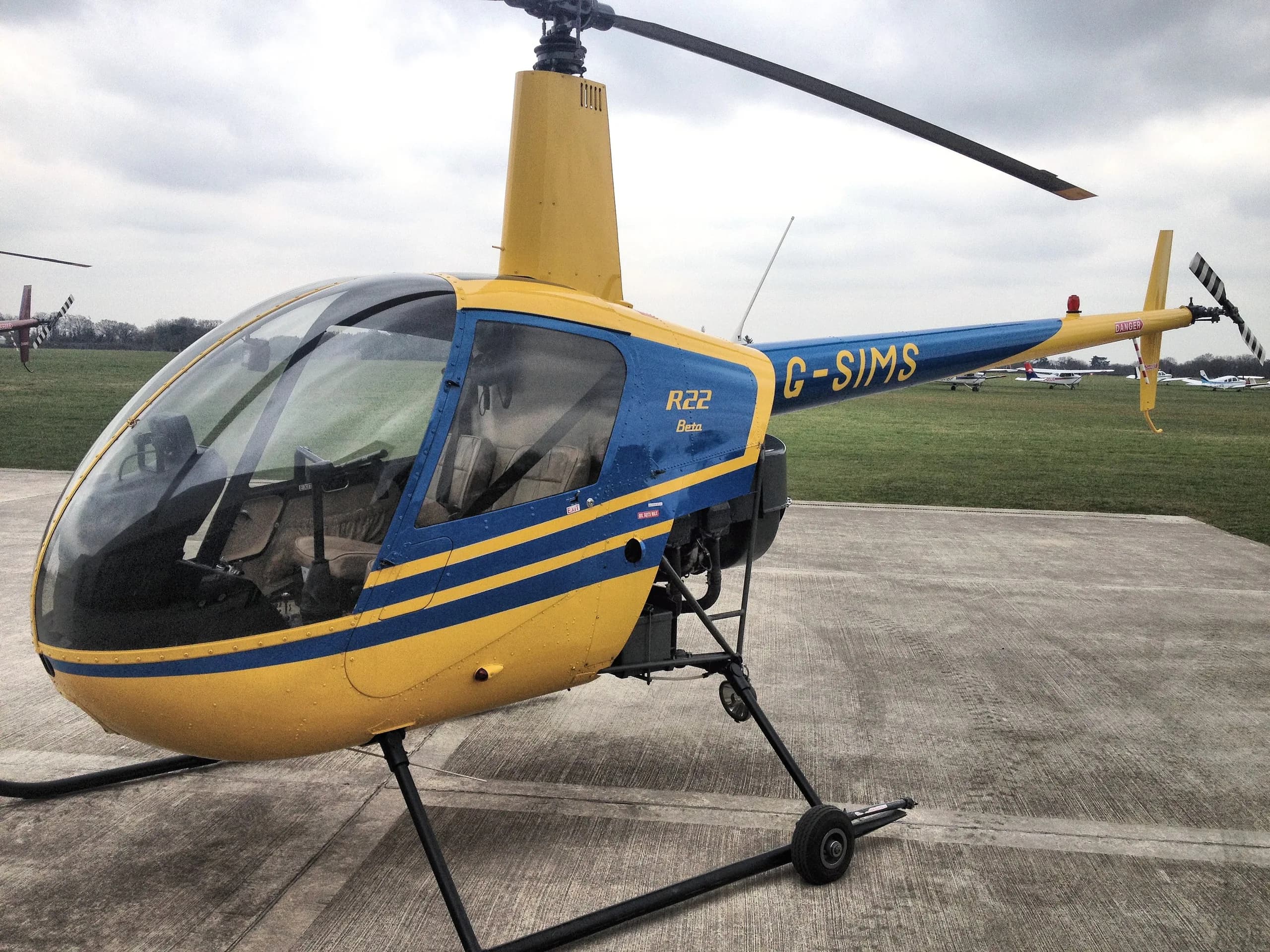 A yellow and blue Robinson R22 Beta helicopter, registration G-SIMS, parked on a tarmac at an airfield.