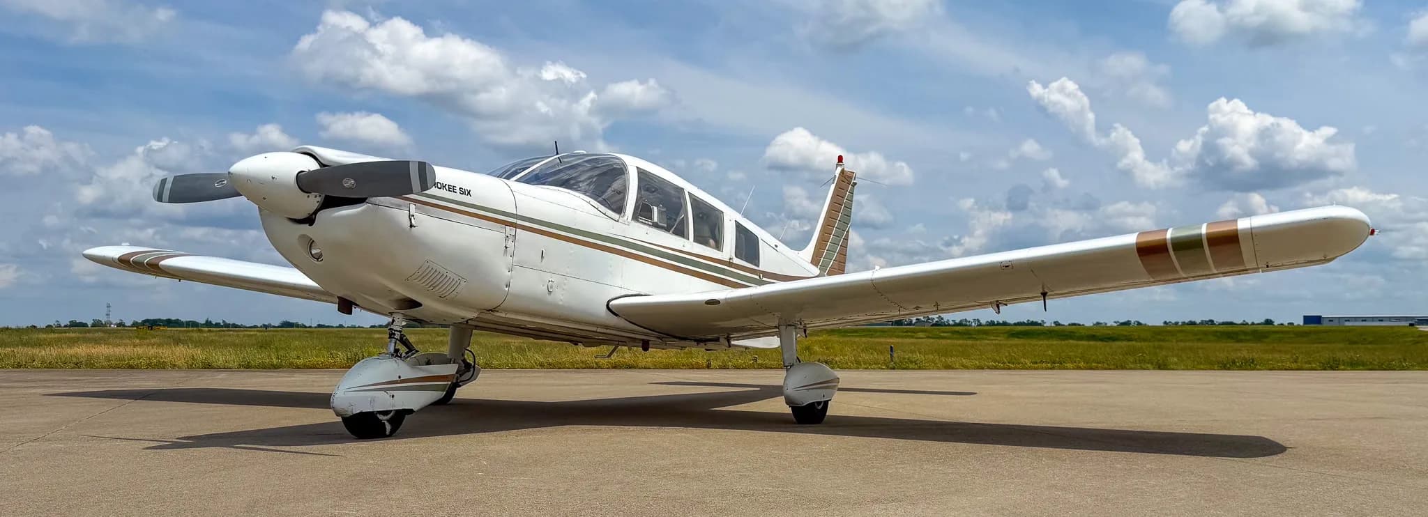 A front-left view of a white Piper Cherokee Six aircraft with brown and green stripes on an airfield under a cloudy sky.
