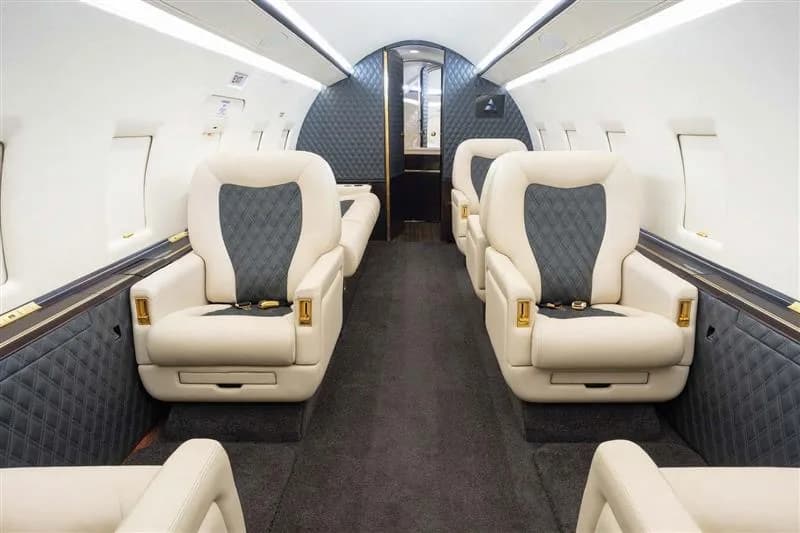 The interior of a private jet cabin showing rows of cream and dark grey quilted seats.