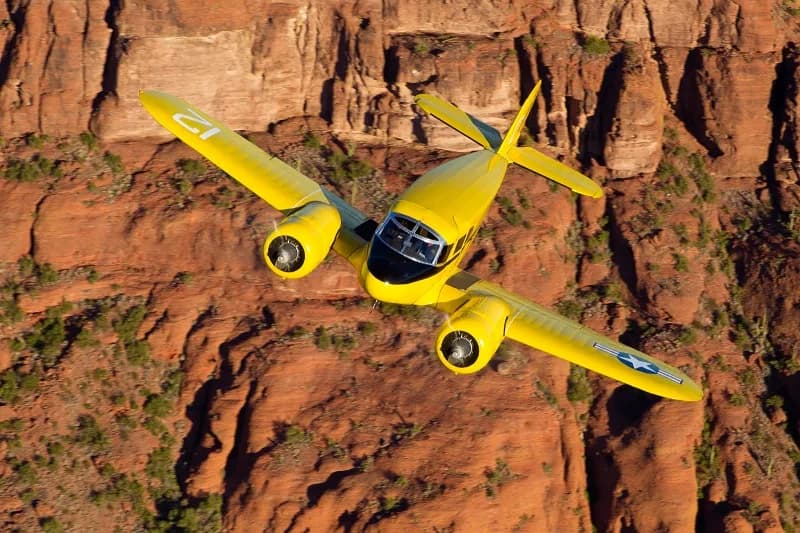A yellow Beechcraft Model 18 flying over a desert landscape.
