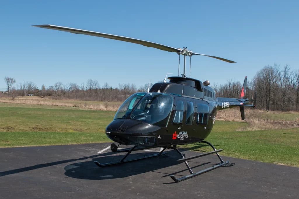 A black Bell 206 helicopter with registration C-GVHD sits on a tarmac with a grassy field and trees in the background.