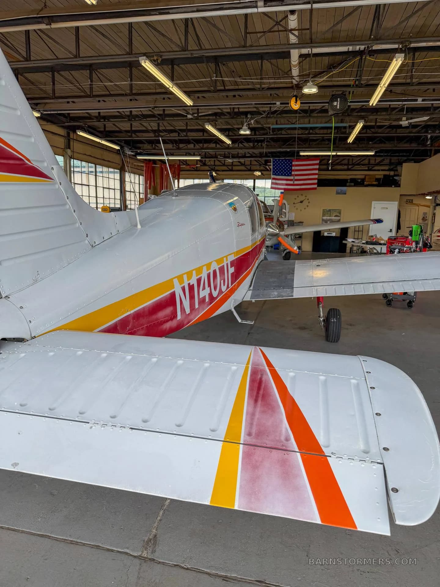 The tail and right wing of a white aircraft with red and yellow stripes, parked inside a hangar.