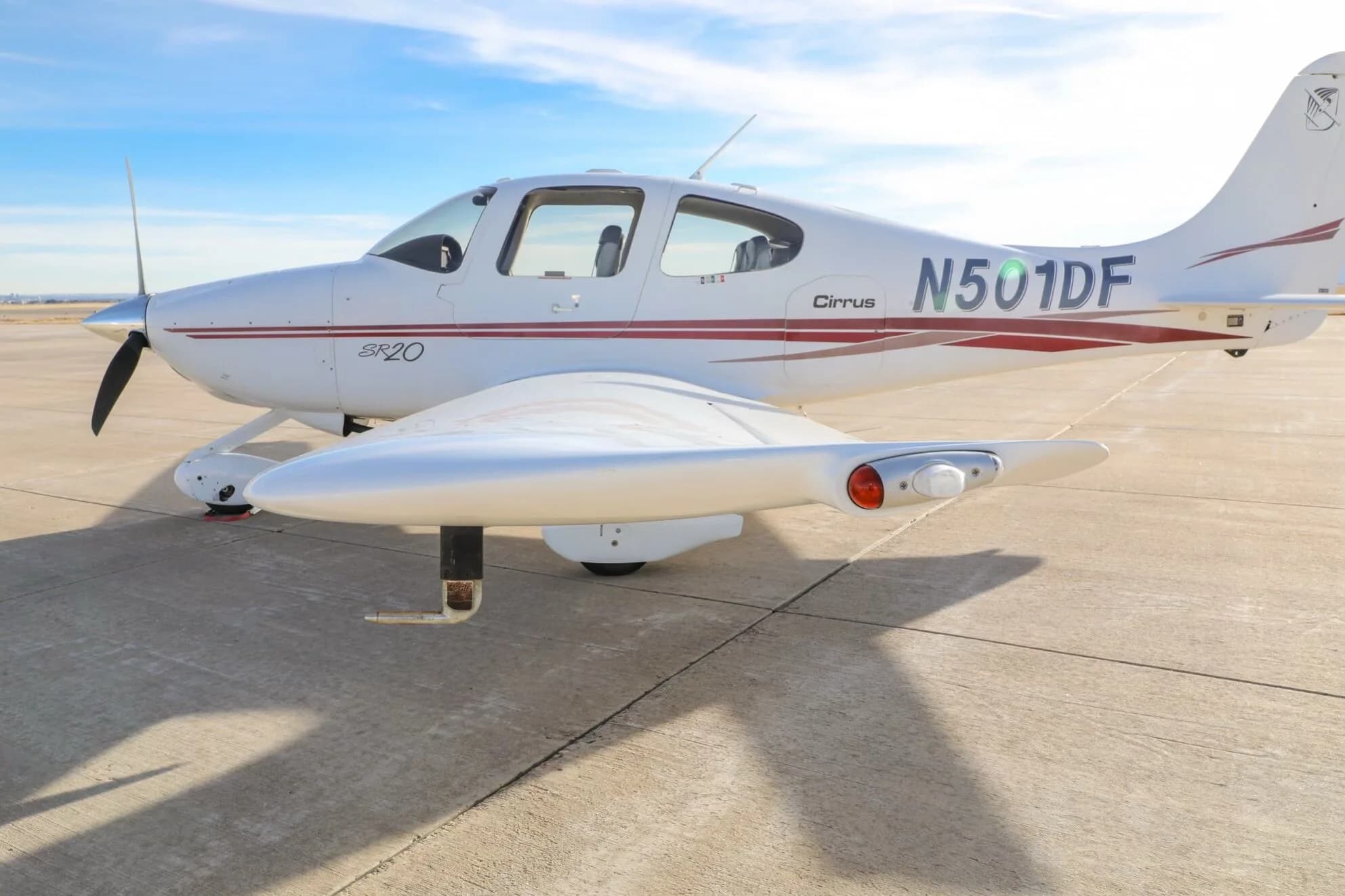 A white Cirrus SR20 aircraft with red stripes, tail number N501DF, parked on an airfield with landing lights illuminated.