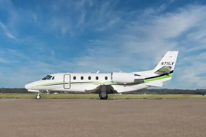 A white business jet with green and black stripes on the tail and fuselage, seen from the side on an airfield under a partly cloudy sky.