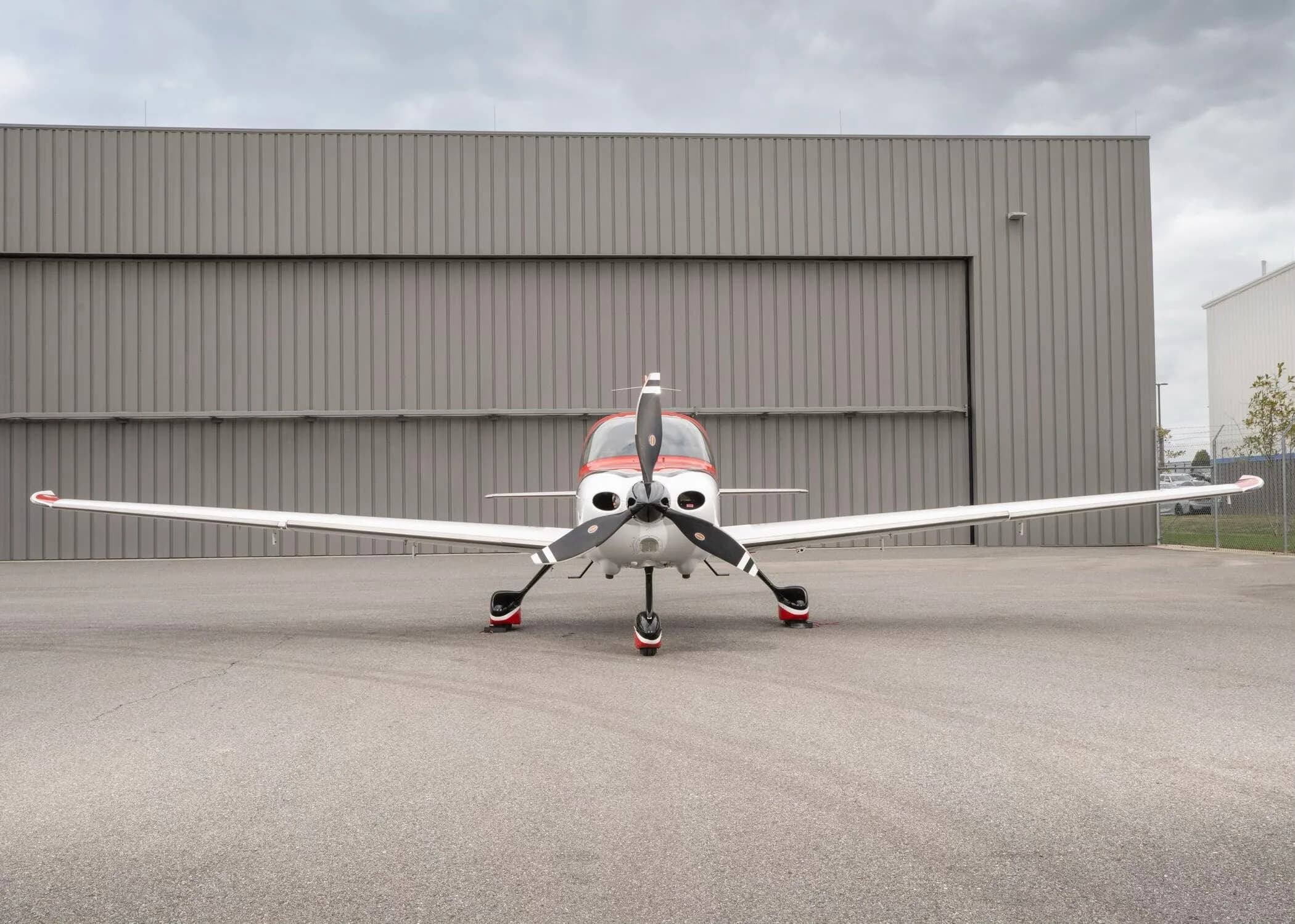 Front view of a red and white Cirrus SR22 aircraft with a three-bladed propeller, parked in front of a hangar.