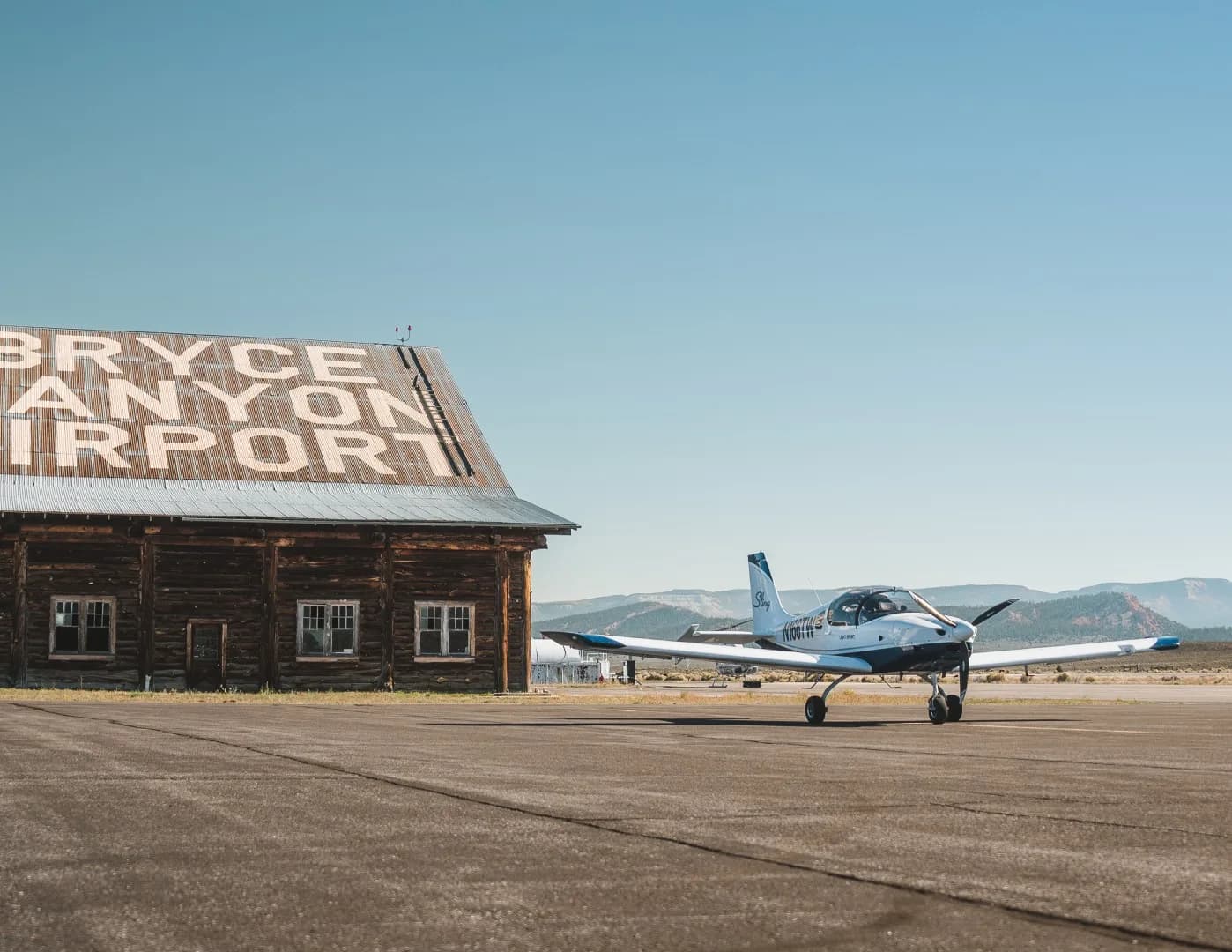 A white and blue Sling Light-Sport aircraft, N166TW, parked on a runway next to a rustic building at Bryce Canyon Airport.