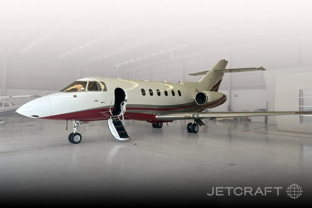 A white and red business jet with its main door open and stairs extended, parked inside a hangar with a faded background.