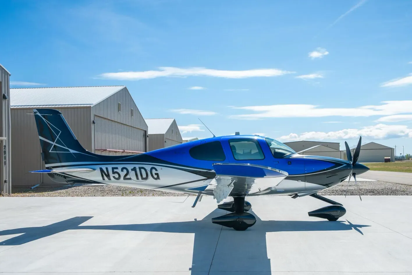 A side view of the blue, black, and white Cirrus SR22T aircraft N521DG parked on a tarmac next to hangars.