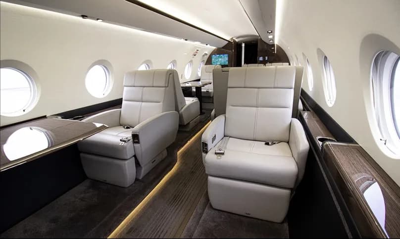 A side view of the private jet cabin showing two rows of light leather seats and large oval windows.