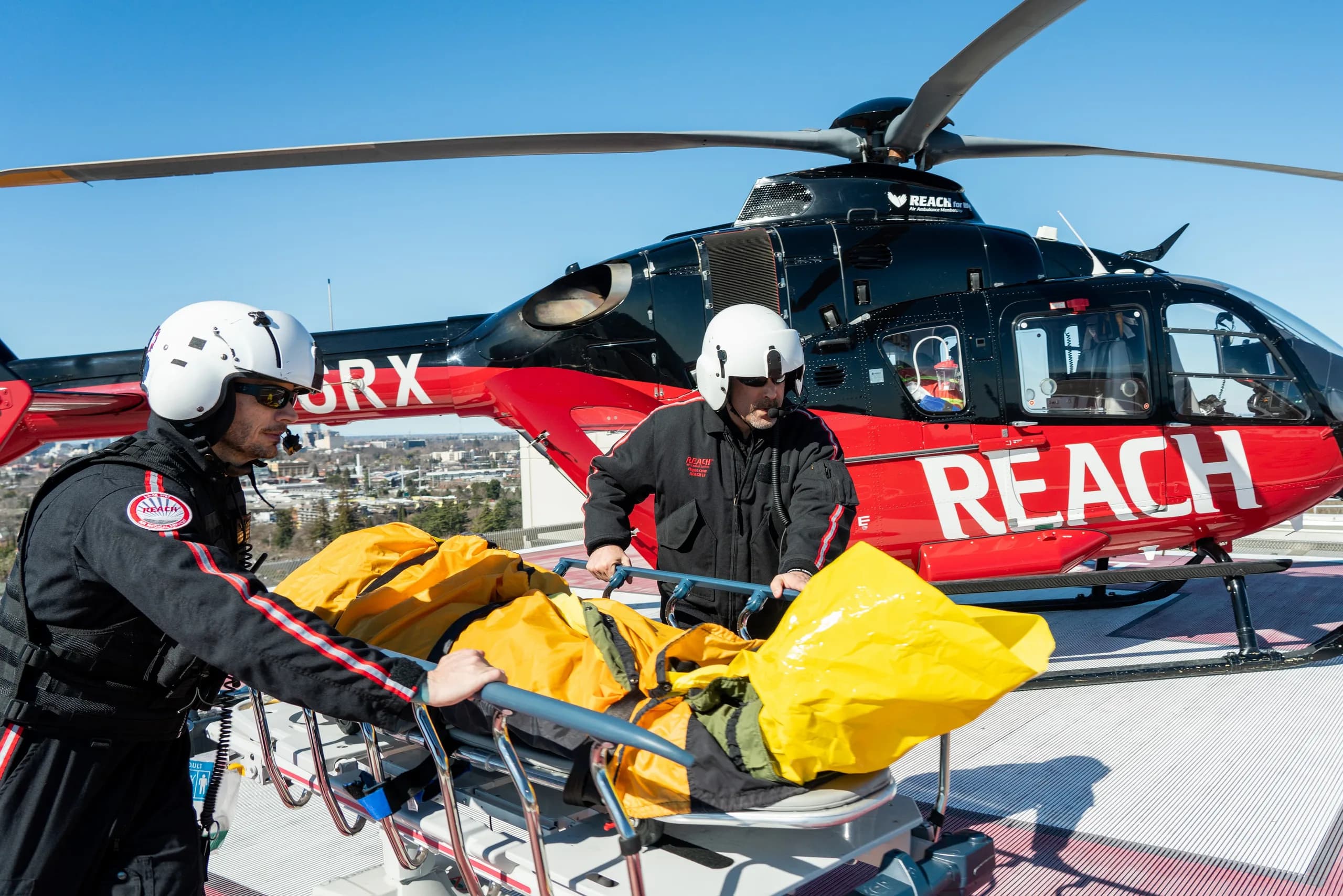 A red and black Airbus EC135 helicopter with 'REACH' markings and medical personnel loading a stretcher.