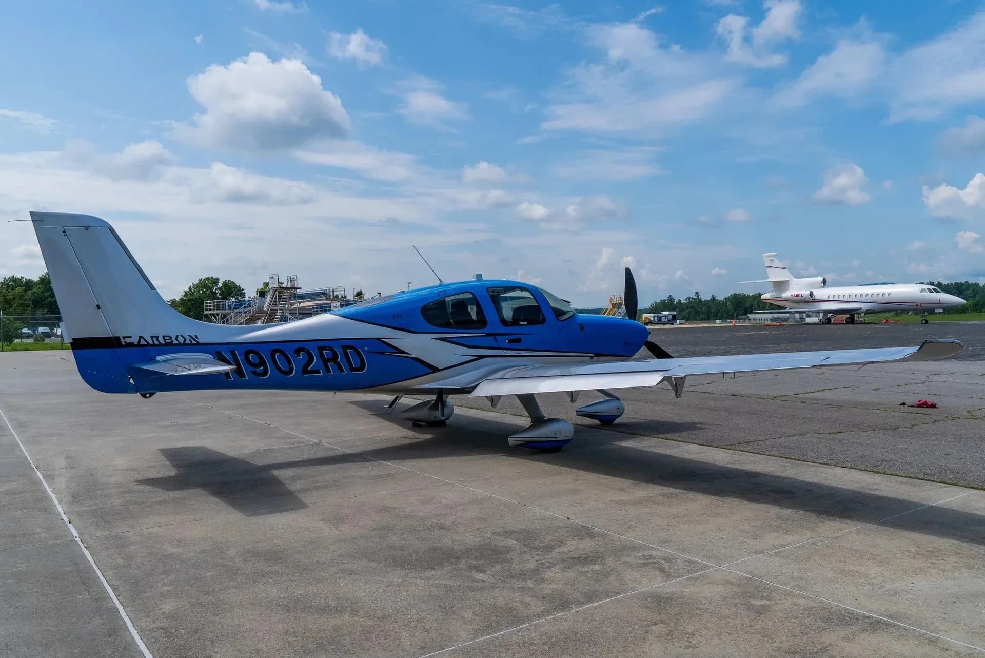 A blue and silver Cirrus SR22 aircraft parked on an airfield with another jet aircraft in the background.