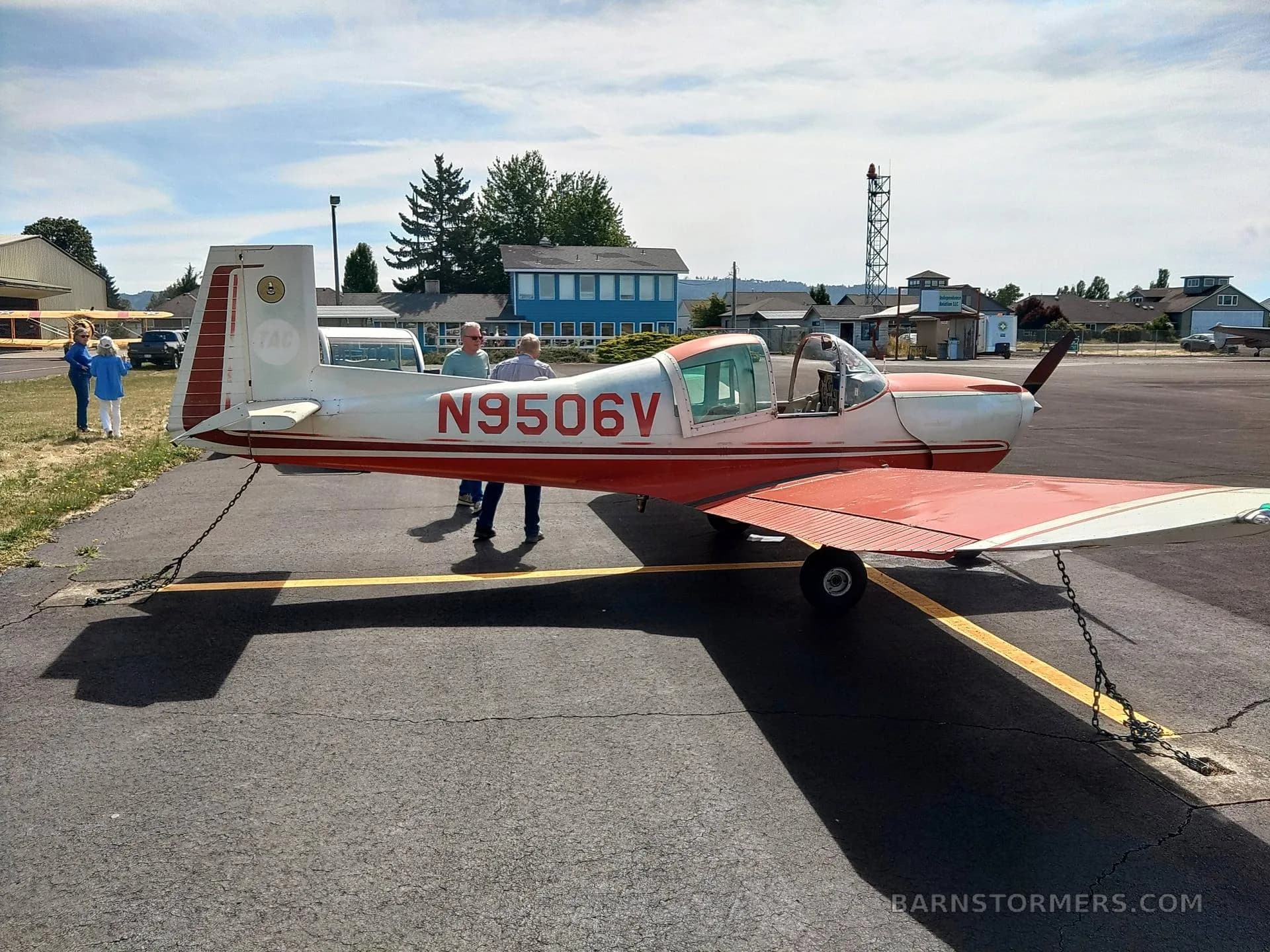 A red and white single-engine aircraft, N9506V, is parked on the tarmac with people standing nearby.