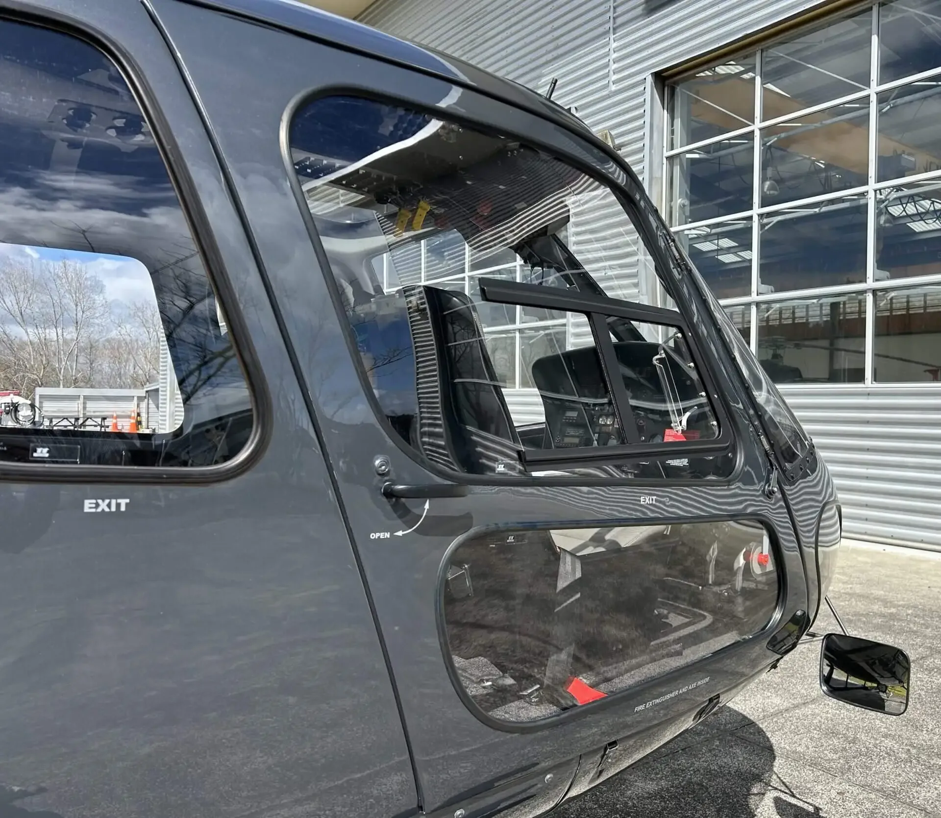 A close-up view of the front section of a dark grey helicopter, showing the cockpit and cabin doors, parked near a hangar.