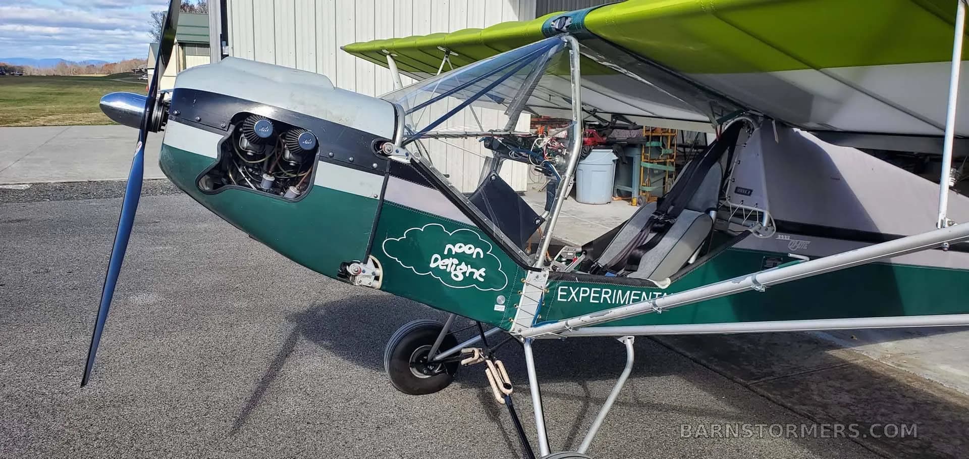 A close-up view of the front-left side of the green and white experimental aircraft, highlighting the "noon delight" cloud graphic and open cockpit.