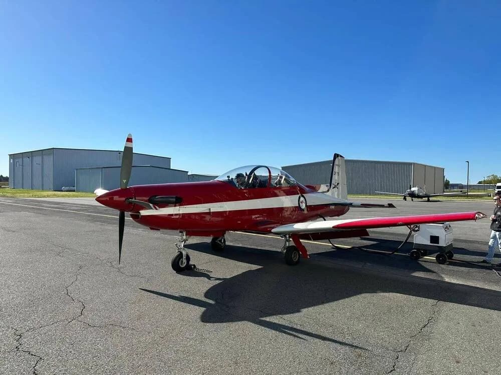 A red and white single-engine aircraft with a bubble canopy is parked on an asphalt tarmac next to hangars.