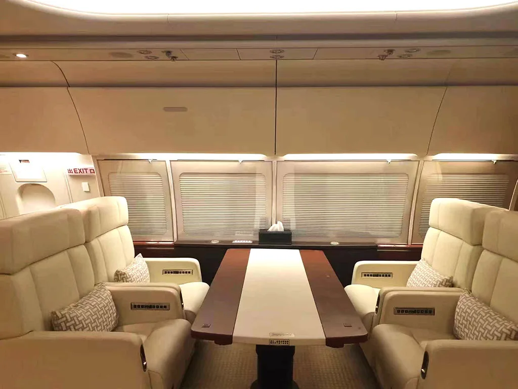 A private jet cabin section with four beige leather seats facing a large wooden table, with window blinds closed.
